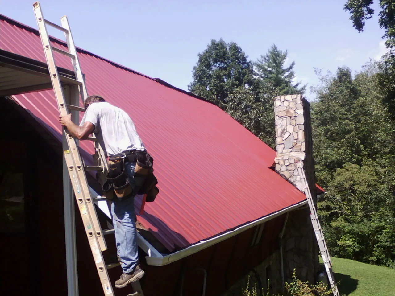 Roofer on a ladder beside a red metal roof and stone chimney — Soffit & Fascia Repair by Roofing Brothers in Mount Union